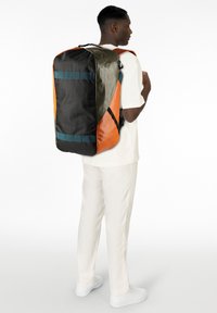 Mochila duffel grande en negro, naranja y verde oliva. Cuenta con correas acolchadas, una textura suave y acentos horizontales en azul.