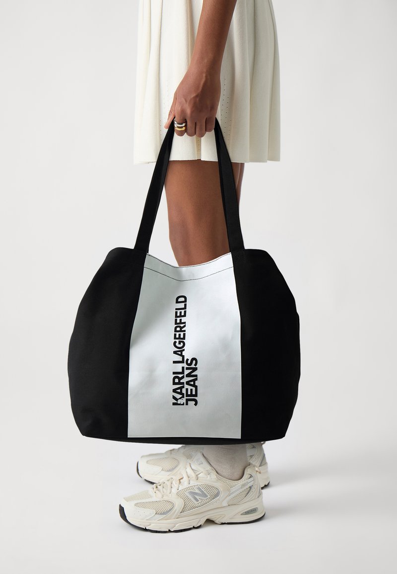 Black and white tote bag with logo, featuring a wide shape, large handles, and fabric material. Visible detail: bold text on side.