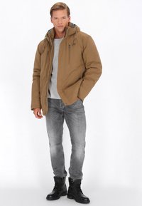 Brun polstret jakke med en aftagelig hætte, der har lynlåse og snore detaljer, iført over en lys grå skjorte og grå jeans med sorte støvler.