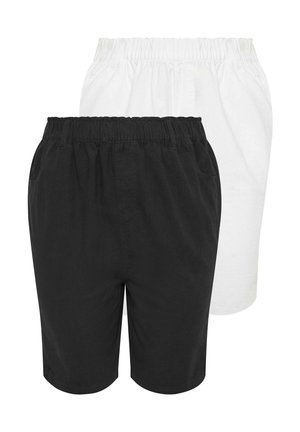 Kobieta w zielonym topie bez rękawów, czarnych krótkich spodniach o wysokim stanie sięgających do kolan oraz czarnych klapkach, stojąca na białym tle.