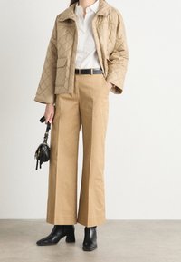 Beige gewatteerde jas met drukknoopsluiting, witte blouse, wijde beige broek en zwarte enkelboots. Zwarte handtas met gouden accenten in de hand.