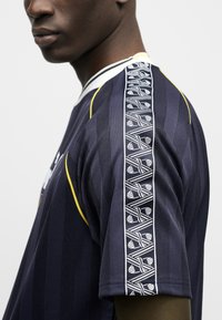 Εγγύς πλάνο ατόμου που φορά μπλε σκούρο πουκάμισο με κάθετο ριγέ μοτίβο και λευκό γεωμετρικό σχέδιο κατά μήκος του μανικιού.