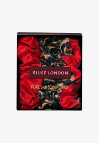 Puhdas silkkinen skrunchie punaisena ja leopardikuosisena, esillä mustassa laatikossa. Tekstissä lukee "SILKE LONDON" ja "PUHDAS SILKKI SCRUNCHIE."