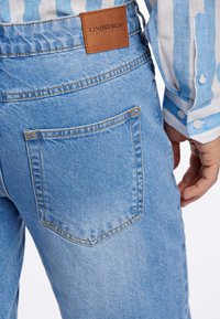 Vaqueros de denim azul claro con bolsillo trasero y etiqueta de marca de cuero marrón. Costuras contrastantes visibles y textura suave.