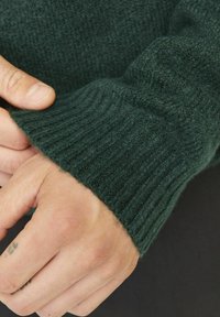 Dunkelgrüner gestrickter Pullover mit gerippten Bündchen, der eine weiche Textur und eine elastische Passform zeigt. Nahaufnahme einer Hand, die das Ärmel-Detail hält.