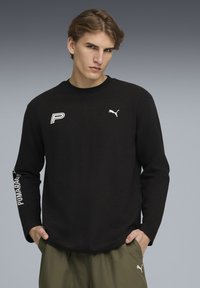 Czarna koszula z długim rękawem o strukturalnym materiale, z białymi logo Puma i "P" na piersi oraz rękawie. W komplecie z oliwkowo-zielonymi spodniami.