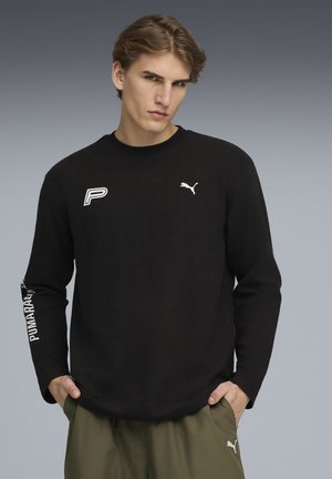 Schwarzes Langarmshirt aus strukturiertem Stoff, mit weißen Puma- und "P"-Logos auf der Brust und dem Ärmeln. Kombiniert mit olivgrünen Hosen.