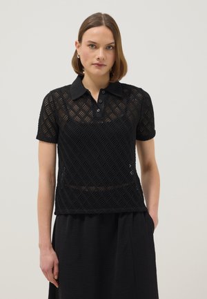 Vrouw met kort bruin haar, gekleed in een zwart, kortmouwig gebreid poloshirt en een zwarte rok, staand met één hand in de zak tegen een effen achtergrond.