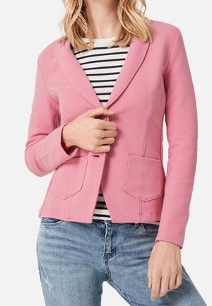 Kvinde iført en pink blazer over en sort-hvidstribet skjorte og blå jeans, holder blazeren lukket med én hånd.