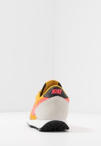 Nike-sneaker med en gul och grå överdel, svart hälkappa, rosa accenter och texturerat mockamaterial på hälen. Platt gummisula.