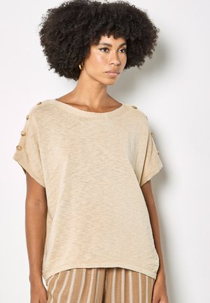 Mujer con cabello rizado, vistiendo una blusa beige de manga corta con botones en las mangas y pantalones marrones con rayas, de pie contra una pared lisa.