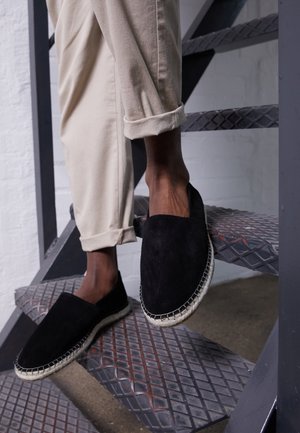 Espadrilles en daim noir avec une semelle en jute tissée, posées sur un escalier en métal texturé, associées à un pantalon khaki retroussé.