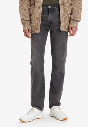 Persoon draagt zwarte vervaagde jeans met rechte pijpen, een bruine gebreide cardigan en witte sneakers met gumzolen, staand tegen een witte achtergrond.