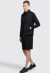 Sort puffervest med høj krave og patchlommer, parret med sorte shorts og sorte sneakers. Slank, moderne design med en mat overflade.