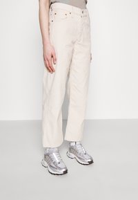 Pantalon large en velours côtelé beige clair avec cinq poches, doté de rivets en cuivre et d'une fermeture à bouton. Associé à des baskets argentées métalliques.