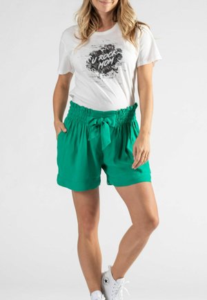 T-shirt blanche avec le graphisme "U ROCK MOM", associé à un short vert à taille élastique avec une ceinture à nouer et des poches, porté avec des baskets blanches.