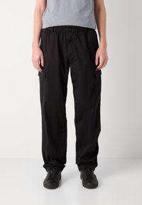 Pantalons cargo noirs avec une coupe décontractée, une taille élastique, des poches latérales et une texture de tissu lisse. Portés avec un t-shirt gris ajusté.