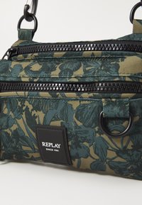 Grün-beige Tasche mit Blumenmuster aus strukturiertem Stoff; verfügt über zwei schwarze Reißverschlüsse und ein rechteckiges Logo-Patch.