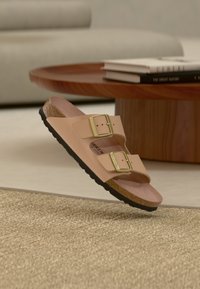 Ταμπά σανδάλι με δύο λουριά και χρυσές αγκράφες που αιωρείται πάνω από μια υφασμάτινη μπεζ κουρελού, κοντά σε ένα ξύλινο τραπεζάκι γεμάτο βιβλία και φόντο έναν καναπέ.