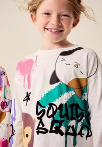Vit långärmad skjorta med färgglada djurgrafik, färgstänk och texten "SQUAD." Materialet verkar vara bomullsliknande med en mjuk yta.