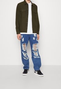 Oliwkowo-zielona koszula-żakiet z zapięciem na guziki z przodu i dwoma kieszeniami na piersi, w zestawieniu z podartymi niebieskimi jeansami z przetarciami i postrzępionymi krawędziami.