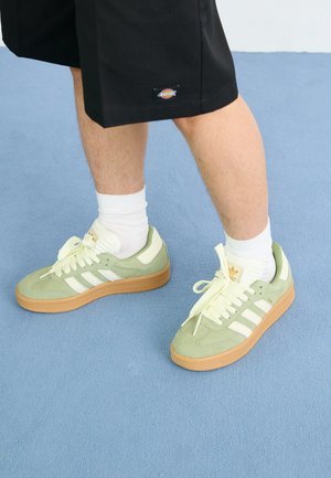 Light green suede sneakers with white stripes and laces, gum sole, and padded collar, paired with white socks and black shorts.