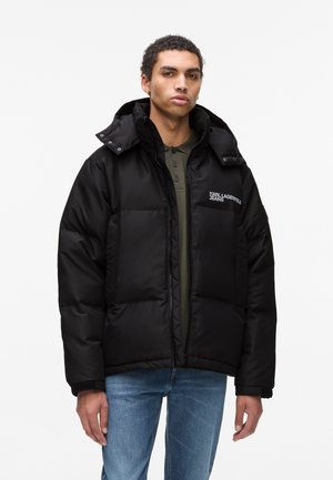 Black puffer jacket with a hood, featuring quilted sections, front zipper, and logo detail on the left chest. Worn over a green top.
