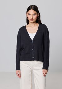 Ciemnoniebieski sweter z dzianiny z dekoltem w serek, sze�ścioma guzikami i ściągaczami, zestawiony z białą bluzką i jasnymi spodniami.