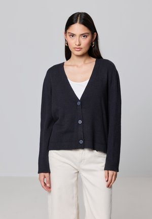 Cardigan tricoté bleu marine foncé avec un col en V, six boutons et des poignets à côtes, associé à un haut blanc et à un pantalon clair.