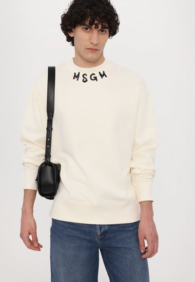 Sweatshirt crème avec le logo "MSGM" noir au niveau du col, associé à un jean bleu. Sac bandoulière noir porté sur l'épaule.