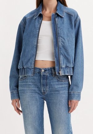 Vrouw draagt een blauwe spijkerjas, gedeeltelijk dichtgeritst over een wit cropped hemdje en blauwe spijkerbroeken tegen een effen achtergrond.
