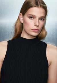 Top sans manches noir à côtes avec un col haut et un design texturé, mettant en valeur des motifs géométriques sur le tissu.