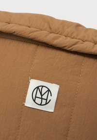 Καφέ τετράγωνο ύφασμα με μια λευκή τετράγωνη ετικέτα ραμμένη πάνω του, που έχει ένα μαύρο κυκλικό λογότυπο από επικάλυψη γραμμάτων.