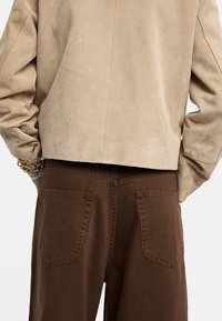 Jasno-beżowa kurtka z zamszu o kroju cropped, z widocznymi detalami szwów, w połączeniu z ciemnobrązowymi spodniami z dwoma tylnymi kieszeniami.