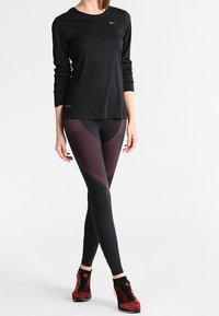 Maglietta sportiva nera a maniche lunghe con un design attillato, abbinata a leggings neri con motivo bordeaux. Presenta un tessuto testurizzato e un'accenno di logo.