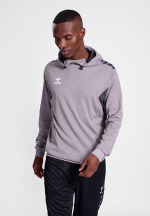 Mand iført lysegrå hættetrøje og sorte træningsbukser, kigger ned og går mod en ensartet baggrund.