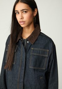 Tumman indigonvärinen denimjacka med en krage i brun kordflossa. Har två framfickor, gula sömmar och dragkedjestängning. Klassisk design.
