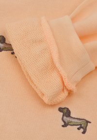 Sweter w kolorze brzoskwiniowym z teksturowanymi mankietami i haftowanym wzorem psa. Materiał jest gładki, z prążkowanymi detalami na mankietach.