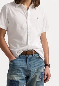 Vit kortärmad skjorta med knappar i en texturerad tyg och liten blå logotyp. Matcher med slitna blå jeans och ett brunt bälte.