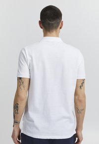 Hvit polo skjorte laget av pustende stoff, med en klassisk krage, kort ermer og rett nederkant. Synlige tatoveringer på armen.
