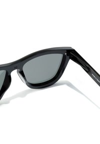 Ochelari de soare negri cu un finisaj lucios, design unghiular, lentile gri, și brațe negre cu textul "Fabricat în Spania".