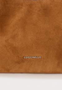 Superficie in pelle scamosciata marrone con logo in metallo argentato "COCCINELLE" centrato vicino al bordo inferiore.