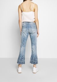 Jean en denim bleu clair avec ourlet évasé, présentant une broderie florale au bas. Conçu avec cinq poches et un aspect usé.