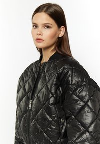 Sort quiltet bomberjakke med en blank tekstur, der har en frontlynlås og ribbede manchetter. Stoffet har et diamantmønster.