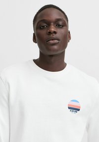 Witte sweatshirt met een rond logo met blauwe, roze en oranje strepen. Zachte textuur en klassiek crew neck ontwerp.