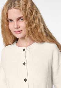 Cremefarbene Strickjacke mit Rundhalsausschnitt, ausgestattet mit fünf dunklen Knöpfen und ribbeliger Struktur am Kragen und den Kanten.