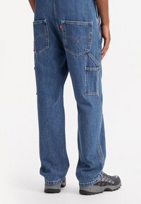 Salopette en denim bleu clair avec jambes droites, deux poches arrière et poches utilitaires sur les côtés. Associée à des chaussures sombres avec des semelles texturées.