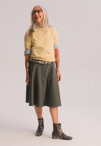 Gul stickad tröja, grå utsvängd kjol, mönstrade svartvita ormskinnsankelboots. Långt hår och glasögon, med synliga tatueringar.