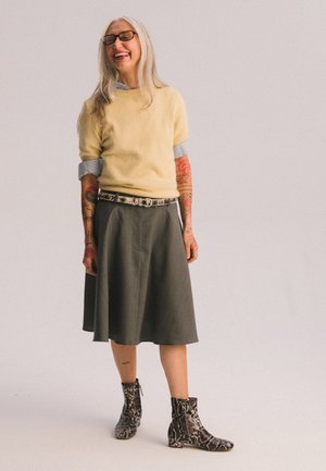 Maglione giallo lavorato a maglia, gonna svasata grigia, stivaletti alla caviglia stampati in bianco e nero con motivo a serpente. Capelli lunghi e occhiali, con tatuaggi visibili.