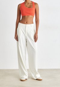 Kobieta w pomarańczowym topie na krótko, szerokich białych spodniach i białych butach, stojąca na tle jednolitego białego tła.